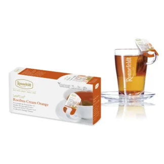 Opakowanie herbaty Ronnefeldt LeafCup Rooibos Cream Orange obok szklanki z przygotowaną herbatą. Herbata Rooibos Cream Orange to czarna herbata z mieszanką przypraw, oferowana w eleganckim pudełku z 15 torebkami herbaty, idealna do przygotowania aromatycznego napoju. Obok znajduje się przezroczysta szklanka z logo Ronnefeldt, ukazująca napój o bogatej, ciemnej barwie.
