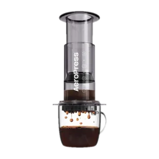 Zdjęcie czarnego zaparzacza AeroPress Clear Black, umieszczonego na szklance z zaparzaną kawą. Krople kawy spadają do kubka, ukazując proces parzenia metodą AeroPress, z przezroczystym czarnym korpusem urządzenia.