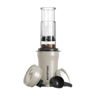 Zdjęcie kremowego ekspresu AeroPress Go Plus, w skład którego wchodzi cylindryczna komora do parzenia kawy, tłok z miarką, pokrywka podróżna oraz miarka do kawy. Urządzenie jest kompaktowe i idealne do przygotowywania kawy w podróży.